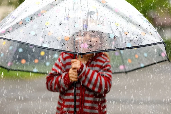 Kleines Mädchen, das an einem regnerischen Tag mit einem großen Regenschirm spielt. Glückliches positives Kind, das durch Regen, Pfützen rennt. Vorschulkind mit Regenkleidung und Gummistiefeln. Kinderaktivität am Schlechtwettertag. — Stockfoto