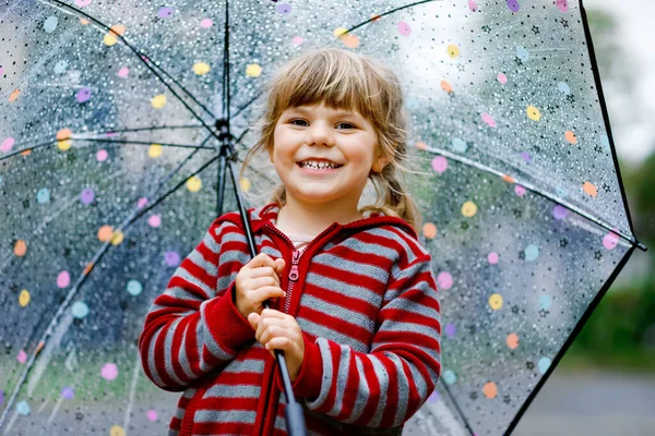 En liten småbarnstjej som leker med ett stort paraply på regnig dag. Lyckligt positivt barn som springer genom regn, pölar. Förskolebarn med regnkläder och gummistövlar. Barnaktivitet på dåligt väder dag. — Stockfoto