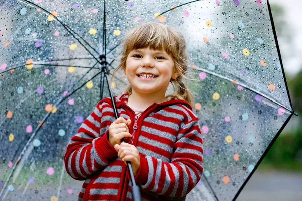 Kleines Mädchen, das an einem regnerischen Tag mit einem großen Regenschirm spielt. Glückliches positives Kind, das durch Regen, Pfützen rennt. Vorschulkind mit Regenkleidung und Gummistiefeln. Kinderaktivität am Schlechtwettertag. — Stockfoto