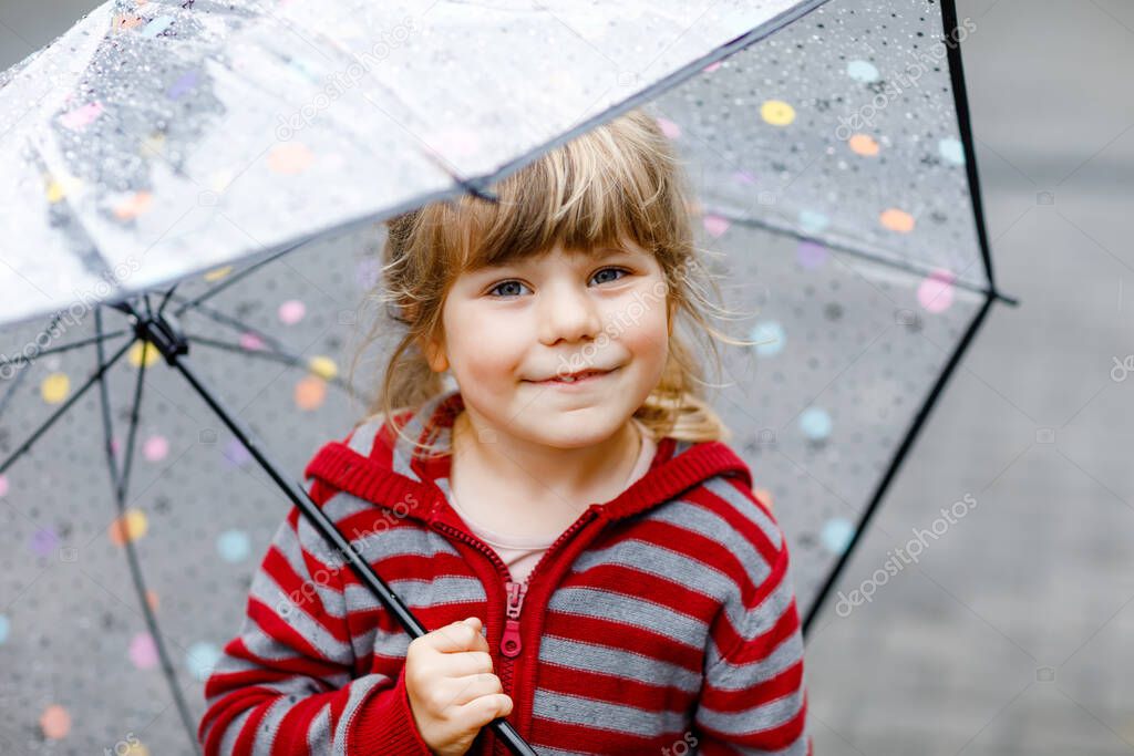 Little toddler girl playing with big umbrella on rainy day. Happy positive child running through rain, puddles. Preschool kid with rain clothes and rubber boots. Children activity on bad weather day.