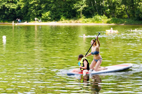 Mujer joven con la chica de la escuela remando en sup board en un lago. Familia activa en moderno moderno stand up paddle board. Actividades de verano al aire libre para familias y niños. Actividades de deportes acuáticos. — Foto de Stock