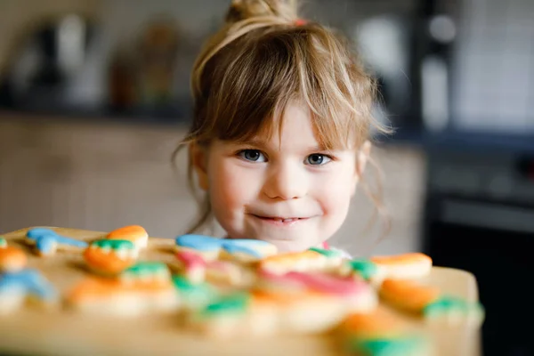 Cute little toddler girl and fresh baked homemade Easter or spring cookies at home indoors. Adorable blond child with apron with bunny and carrot cookie in domestic kitchen. Child eating cookie — Stock Photo, Image