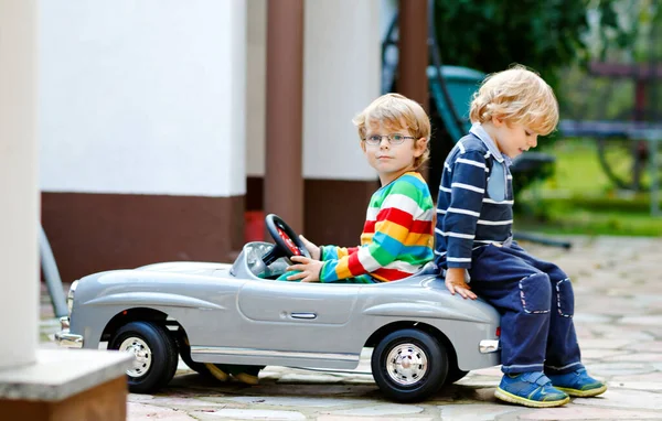 Dva malí školáci si hrají s velkým starým autíčkem v letní zahradě venku. Šťastné děti si spolu hrají, řídí auto. Venkovní aktivita pro děti. Sourozenci a přátelé v teplý den — Stock fotografie