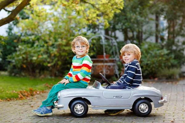 Dos niños preescolares jugando con un gran coche de juguete viejo en el jardín de verano, al aire libre. Los niños felices juegan juntos, conduciendo el coche. Actividad al aire libre para niños. Hermanos y amigos en un día cálido — Foto de Stock