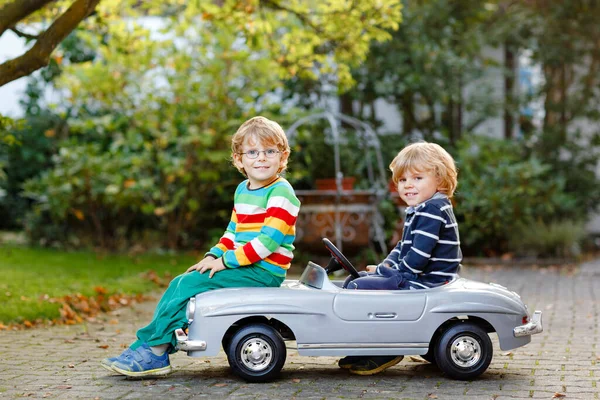 Due bambini in età prescolare che giocano con una grande macchina giocattolo nel giardino estivo, all'aperto. Bambini felici giocano insieme, guidano la macchina. Attività all'aperto per bambini. Fratelli e amici nella giornata calda — Foto Stock