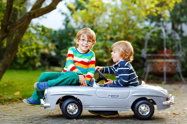 Dwóch małych przedszkolaków bawiących się dużym starym samochodzikiem w letnim ogrodzie, na świeżym powietrzu. Szczęśliwe dzieci bawią się razem, prowadzą samochód. Aktywność na świeżym powietrzu dla dzieci. Rodzeństwo i przyjaciele w ciepły dzień — Zdjęcie stockowe