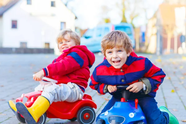 Zwei kleine Jungen in bunten Klamotten und Regenstiefeln fahren Spielzeugautos. Geschwister machen einander Konkurrenz, im Freien. Aktive Freizeit für Kinder am Herbsttag. — Stockfoto