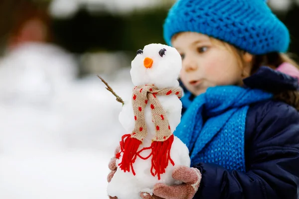 かわいい小さな幼児の女の子はミニ雪だるまを作り、ニンジンの鼻を食べる。寒い日には屋外で雪と遊ぶと楽しい健康的な幸せな子供。冬の子供たちとのアクティブなレジャー — ストック写真