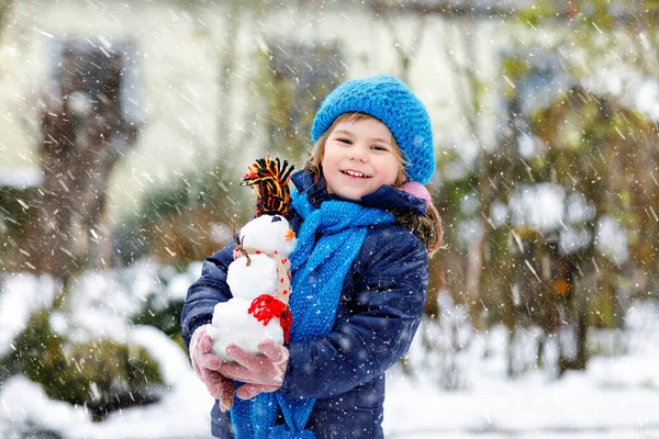 Linda niñita haciendo mini muñeco de nieve y comiendo la nariz de zanahoria. Adorable niño feliz saludable jugando y divirtiéndose con la nieve, al aire libre en el día frío. Ocio activo con niños en invierno —  Fotos de Stock