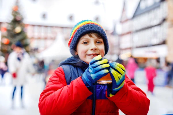 Niño feliz en ropa de abrigo colorido en pista de patinaje del mercado de Navidad o feria beber ponche caliente o chocolate. Niño sano divirtiéndose en patinaje sobre hielo. personas que tienen ocio activo de invierno — Foto de Stock