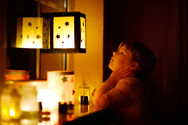 Malá holčička sedící u okna s ručně vyrobenými lucernami se svíčkami pro průvod sv. Martina. Dítě batole se dívá na zářící lucernu. Německá tradice Martinsumzug. Domácí dekorace — Stock fotografie