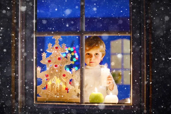 Lindo menino, parado à janela na época do Natal e segurando vela. Criança à espera do Pai Natal. e presentes. Com luzes coloridas da árvore de Natal no fundo — Fotografia de Stock