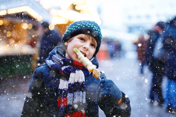 Pequeno menino bonito comendo frutas cobertas de chocolate branco no espeto no tradicional mercado de Natal alemão. Criança feliz no mercado familiar tradicional na Alemanha durante o dia nevado. — Fotografia de Stock