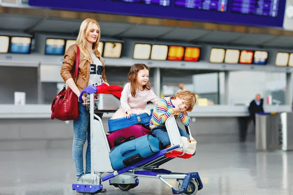 Dos niños pequeños, niño y niña, hermanos y madre en el aeropuerto. Niños, familia viajando, yendo de vacaciones en avión y esperando en el carro con maletas empujando por la mujer en la terminal para el vuelo. —  Fotos de Stock