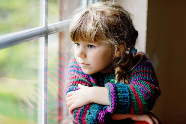 Holčička sedí u okna. Dítě ve školce nosí útulný samočerný vlněný svetr. Batole se dívá venku na déšť. Podzimní a zimní sezóna. — Stock fotografie