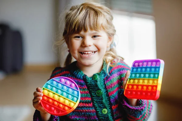 Piccola ragazza prescolare bionda che gioca con il nuovo giocattolo sensoriale di tendenza - arcobaleno pop it. Giocattolo antistress per bambini e adulti. Giocattolo colorato semplice fossetta. Squishy morbido bolla giocattoli arcobaleno colore. Bambino felice. — Foto Stock