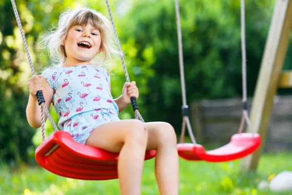 Glad liten småbarn flicka som har kul på swing i hushållsträdgården. Leende positiva friska barn svänger på solig dag. Förskoleflickan skrattar och gråter. Aktiv fritid och aktivitet utomhus. — Stockfoto