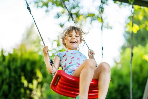Glad liten småbarn flicka som har kul på swing i hushållsträdgården. Leende positiva friska barn svänger på solig dag. Förskoleflickan skrattar och gråter. Aktiv fritid och aktivitet utomhus. — Stockfoto