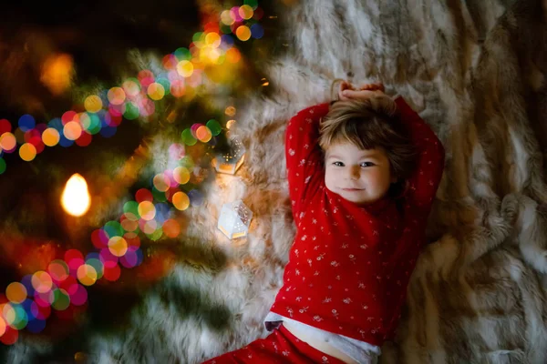 stock image Little cute toddler girl lies under Christmas tree and dreaming of Santa at home, indoors. Traditional Christian festival. Happy kid child waiting for gifts on xmas. Cozy soft light