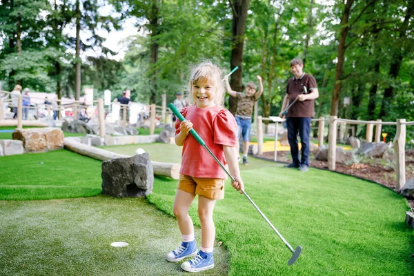 Χαριτωμένο κορίτσι προσχολικής ηλικίας παίζει μίνι γκολφ με την οικογένεια. Ευτυχισμένο μικρό παιδί που διασκεδάζει με την υπαίθρια δραστηριότητα. Θερινό άθλημα για παιδιά και ενήλικες, σε εξωτερικούς χώρους. Οικογενειακές διακοπές ή θέρετρο. — Φωτογραφία Αρχείου