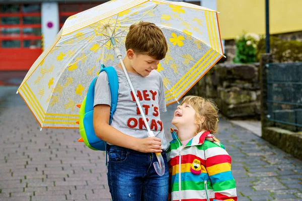 Schulkind Junge holt die kleine Schwester, Vorschulmädchen aus dem Kindergarten ab. Zwei glückliche Kinder mit Regenschirm, die bei Regen spielen und bei schlechtem Wetter Spaß haben. Verliebte Geschwister. — Stockfoto