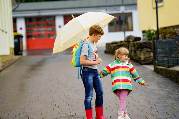 小学生男孩接妹妹，学龄前女孩从幼儿园。两个快乐的孩子带着雨伞在雨天玩耍，在恶劣的雨天玩得很开心。#坠入爱河#. — 图库照片