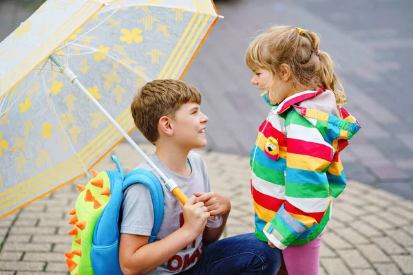 Chłopak ze szkoły odbiera młodszą siostrę, przedszkolankę z przedszkola. Dwoje szczęśliwych dzieci z parasolem bawiącym się podczas deszczu i bawiącym się w deszczowe dni. Rodzeństwo zakochane. — Zdjęcie stockowe