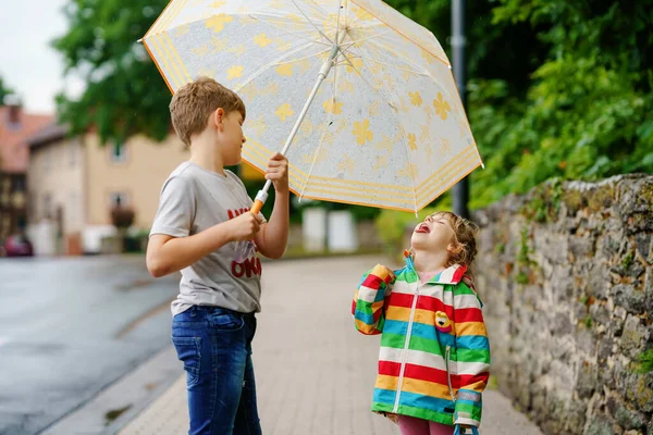 School kid boy picks up the little sister, preschool girl from kindergarten. Two happy children with umbrella playing during rain and having fun on bad weather rainy day. Siblings in love. — Stock Photo, Image