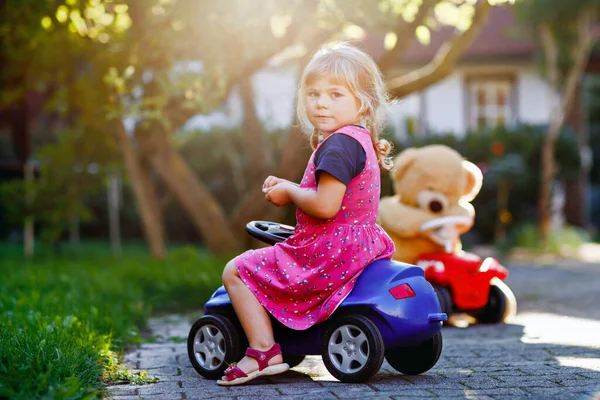 Pequena menina adorável criança dirigindo carro de brinquedo e se divertindo com brincar com urso de brinquedo de pelúcia, ao ar livre. Linda criança saudável feliz desfrutando de dia quente de verão. Sorrindo garoto deslumbrante em gaden — Fotografia de Stock