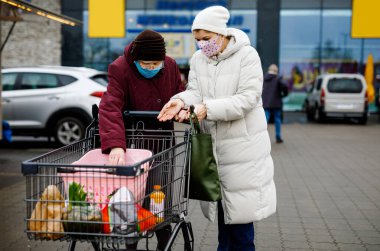 Kıdemli kadın ve tıbbi maskeli sosyal hizmetler görevlisi pandemik koronavirüs hastalığı nedeniyle. Kızı ya da torunu süpermarkette alışveriş yaparken büyükannesine yardım ediyor, market arabasında yiyecek satıyor, açık havada geziyor.