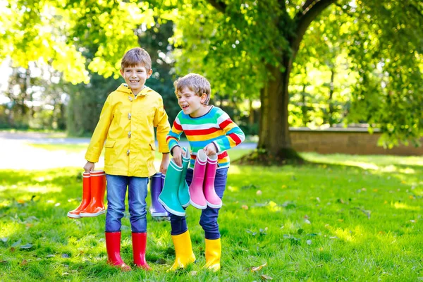 2人の小さな男の子、カラフルな雨のブーツがたくさんあるかわいい兄弟。異なるゴムブーツやジャケットの子供。雨の秋のための履物。健康的な双子と親友が屋外で楽しんでいます — ストック写真