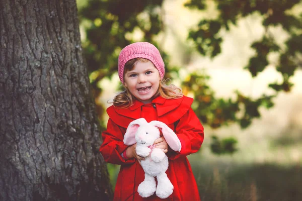 Beautiful toddler girl in red coat making a walk through autumn forest. Happy healthy baby holding soft toy rabbit. Sunny fall day with child. Active leisure and activity with kids in nature. — Stock Photo, Image