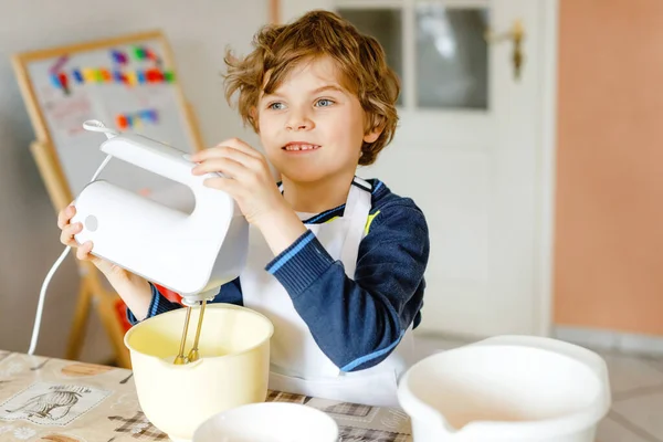 Roztomilý blonďatý chlapeček pečící čokoládový dort a ochutnávající těsto v kuchyni, uvnitř. Šťastné dítě baví s prací s mixérem, moukou, vejce těsto doma. Malý pomocník — Stock fotografie