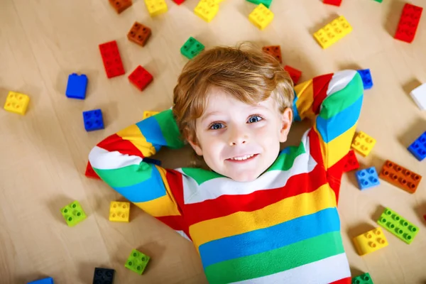 Маленький блондин играет с большим количеством красочных пластиковых блоков в помещении. Мальчик в разноцветной рубашке и веселится со строительством и созданием — стоковое фото