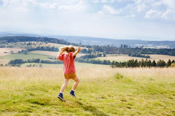 Niña preescolar corriendo y caminando por el prado con flores florecientes en el campo, con vista a las colinas y montañas. Niño feliz divirtiéndose, actividad familiar al aire libre, senderismo en el día de verano. — Foto de Stock