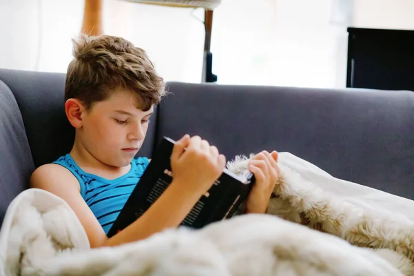 Söt blond liten pojke läser tidningen eller boka i hemmets rum. Spännande barn läser högt, sitter på soffan. Skolbarn, familj, utbildning. Hemundervisning under covid tid — Stockfoto