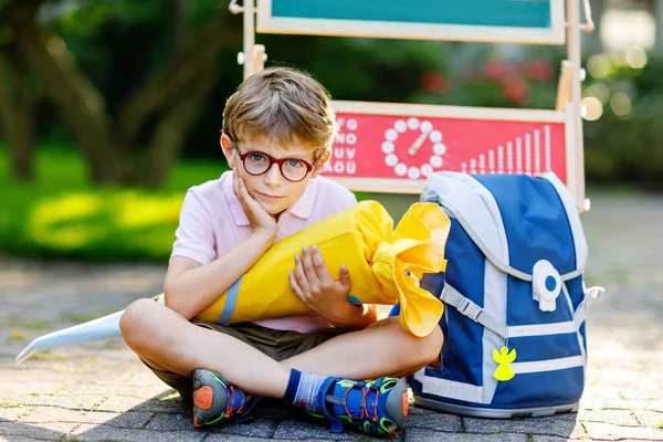 Rapazinho feliz com óculos sentados à secretária e mochila ou mochila. Schoolkid com cone tradicional bolsa de escola alemã chamado Schultuete em seu primeiro dia de escola — Fotografia de Stock