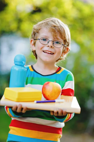 Feliz niño preescolar con libros, manzana y botella de bebida en su primer día a la escuela primaria o guardería. Niño sonriente, estudiante con gafas, al aire libre. Volver al concepto de educación escolar. — Foto de Stock