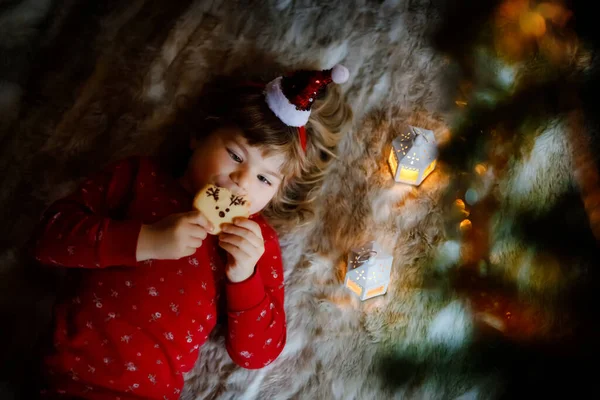Little cute toddler girl in bed under Christmas tree, eating reindeer cookie and dreaming of Santa at home, indoors. Traditional Christian festival. Happy kid child waiting for gifts on xmas. — Stock Photo, Image