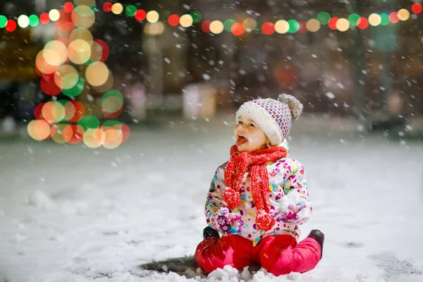Adorable little toddler girl walking outdoors in winter. Cute toddler during strong snowfall on evening. Child having fun with snow. Wearing warm baby colorful clothes and hat with bobbles. — Stock Photo, Image