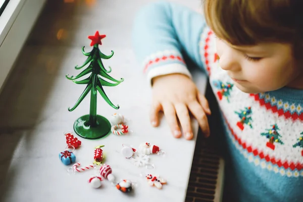 Primer plano de niña por ventana y decorando pequeño árbol de Navidad de vidrio con pequeños juguetes de Navidad. El niño celebra la fiesta familiar tradicional. Primer plano en las manos con el hombre de jengibre — Foto de Stock