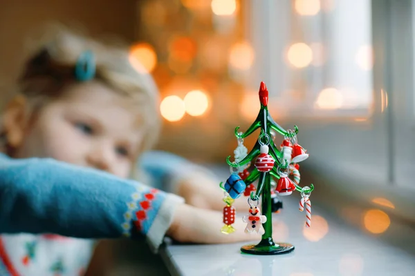 Menina pequena criança sentada à janela e decorando pequena árvore de Natal de vidro com minúsculos brinquedos xmas. A criança sã feliz celebra férias tradicionais de família. Um bebé adorável. Foco seletivo na árvore — Fotografia de Stock