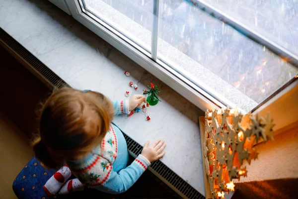 Menina pequena criança sentada à janela e decorando pequena árvore de Natal de vidro com minúsculos brinquedos xmas. A criança sã feliz celebra férias tradicionais de família. Adorável bebê. — Fotografia de Stock
