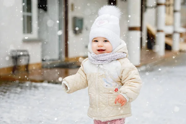 Счастливая маленькая девочка делает первые шаги на улице зимой через снег. Милый малыш учится ходить. Ребенок веселится в холодный снежный день. Первый снег, активность. Зимняя прогулка на природе — стоковое фото