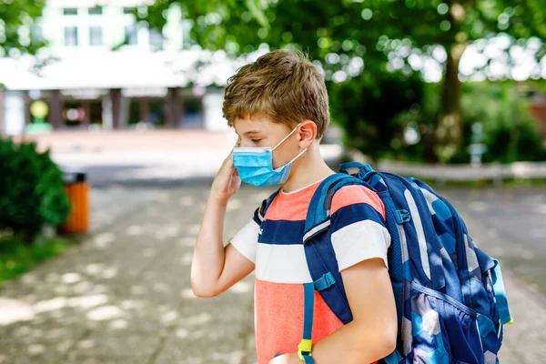 Niño con máscara médica camino a la escuela. Niño con mochila mochila. Volver a la escuela después de las vacaciones de verano. Protección durante la pandemia de corona. Niño sano al aire libre en el patio de la escuela con el edificio — Foto de Stock