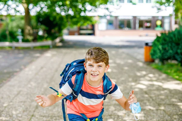 Rapaz com máscara médica a caminho da escola. Criança com mochila mochila. De volta à escola depois das férias de verão. Proteção durante a pandemia de corona. Criança saudável ao ar livre no pátio da escola com edifício — Fotografia de Stock