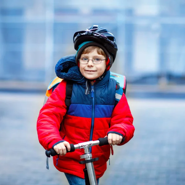 Милый маленький школьник едет на скутере по пути в начальную школу. Ребенок в защитном шлеме, школьная сумка дождливым осенним холодным утром. Движение в городе и школьники. — стоковое фото