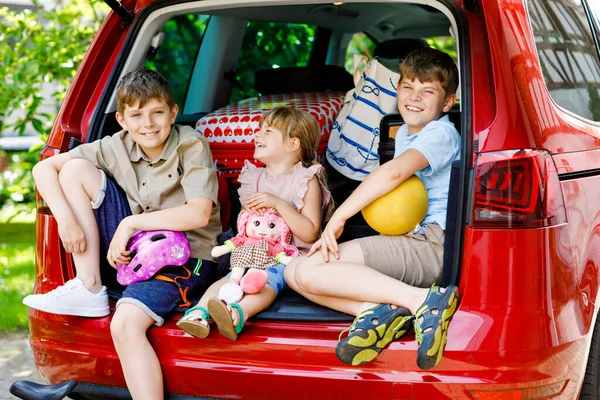 Drie kinderen, twee jongens en een kleuter zitten in de kofferbak voordat ze met hun ouders op zomervakantie gaan. Gelukkige kinderen, broers en zussen, broers en zussen met koffers en speelgoed op reis — Stockfoto