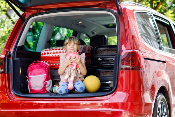 Aranyos kis óvodás lány ül a csomagtartóban, mielőtt elmegy nyaralni a szülőkkel. Boldog aktív gyermek bőröndökkel, kerékpárral és játékokkal családi utazásra, kirándulásra — Stock Fotó