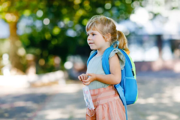 Niña pequeña en su primer día de camino a la escuela de juego con máscara médica contra el virus de la corona covid. Saludable hermoso bebé caminando a la guardería preescolar y jardín de infantes. Niño feliz con mochila —  Fotos de Stock